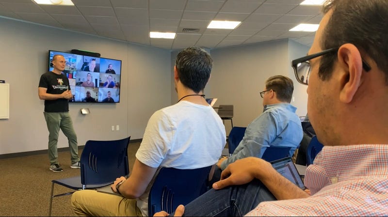 Brett Hurt presents in a conference room to in-person attendees attendees in front of him and a monitor showing remote attendees behind him.